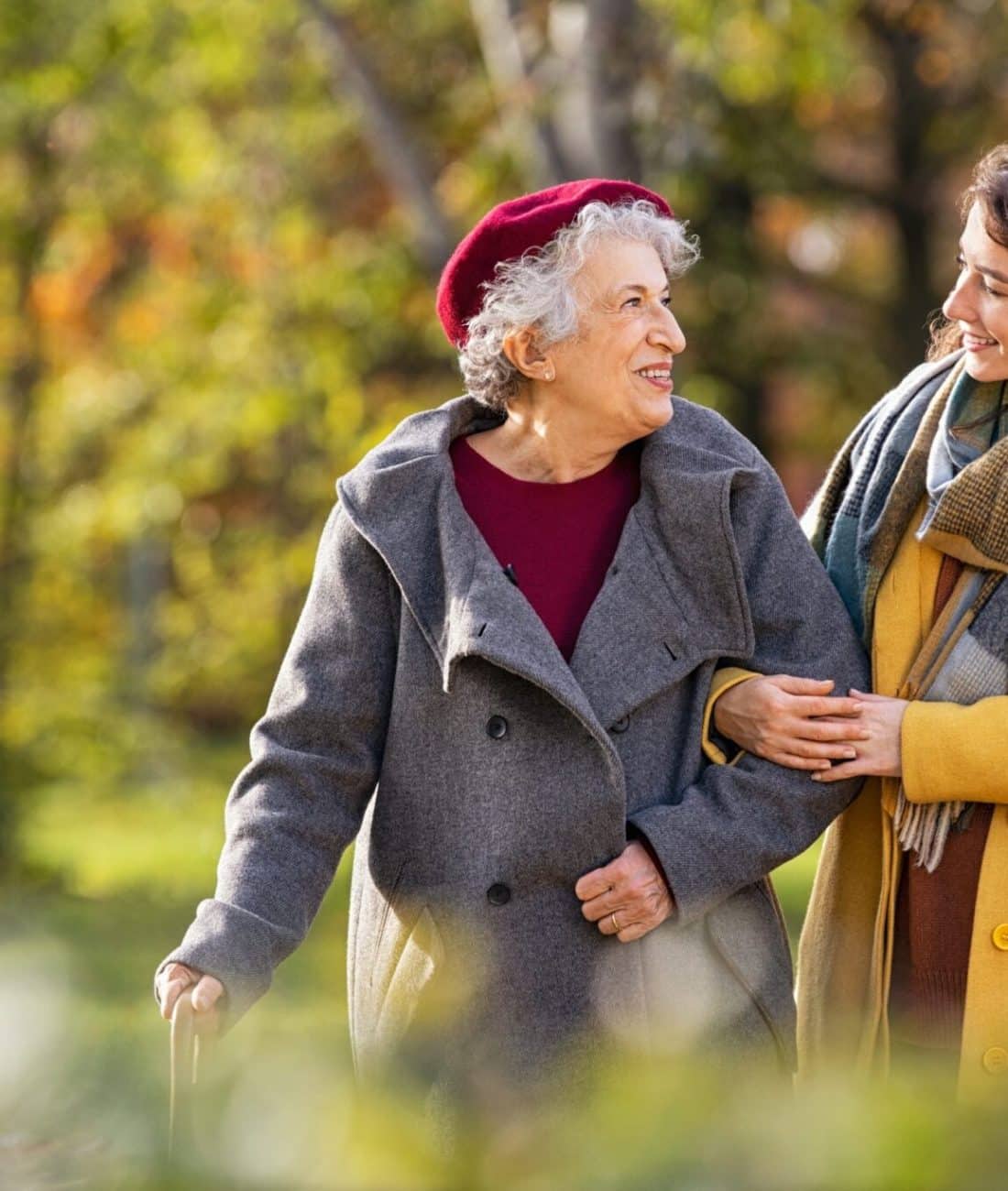 Young woman in park wearing winter clothing walking with old grandmother. Happy grandma wearing coat walking with lovely girl outdoor with copy space. Smiling lovely caregiver and senior lady walking in park during autumn and looking at each other.